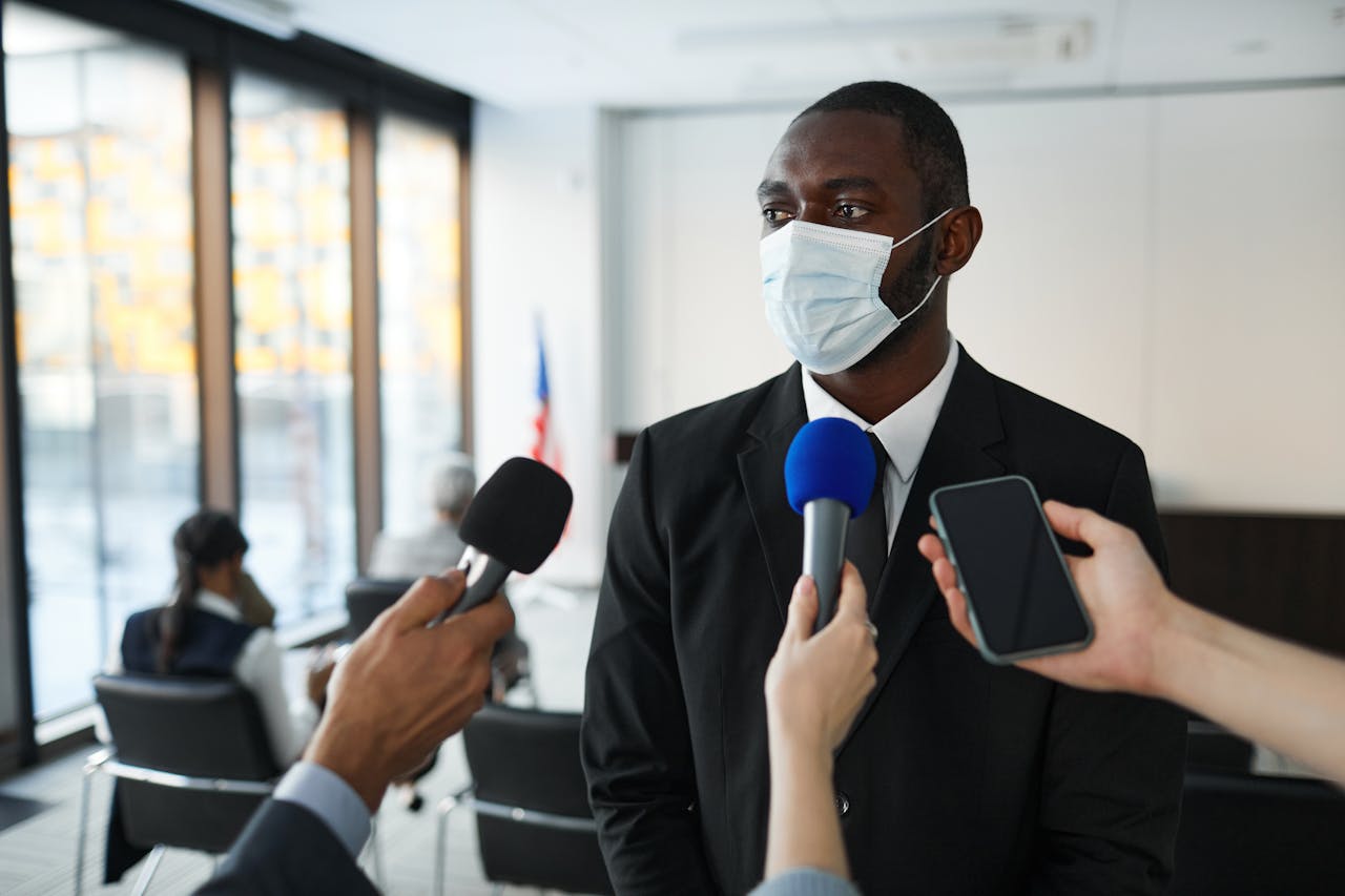services-04 African American businessman in formal attire speaks to media while wearing a face mask in a conference room setting.