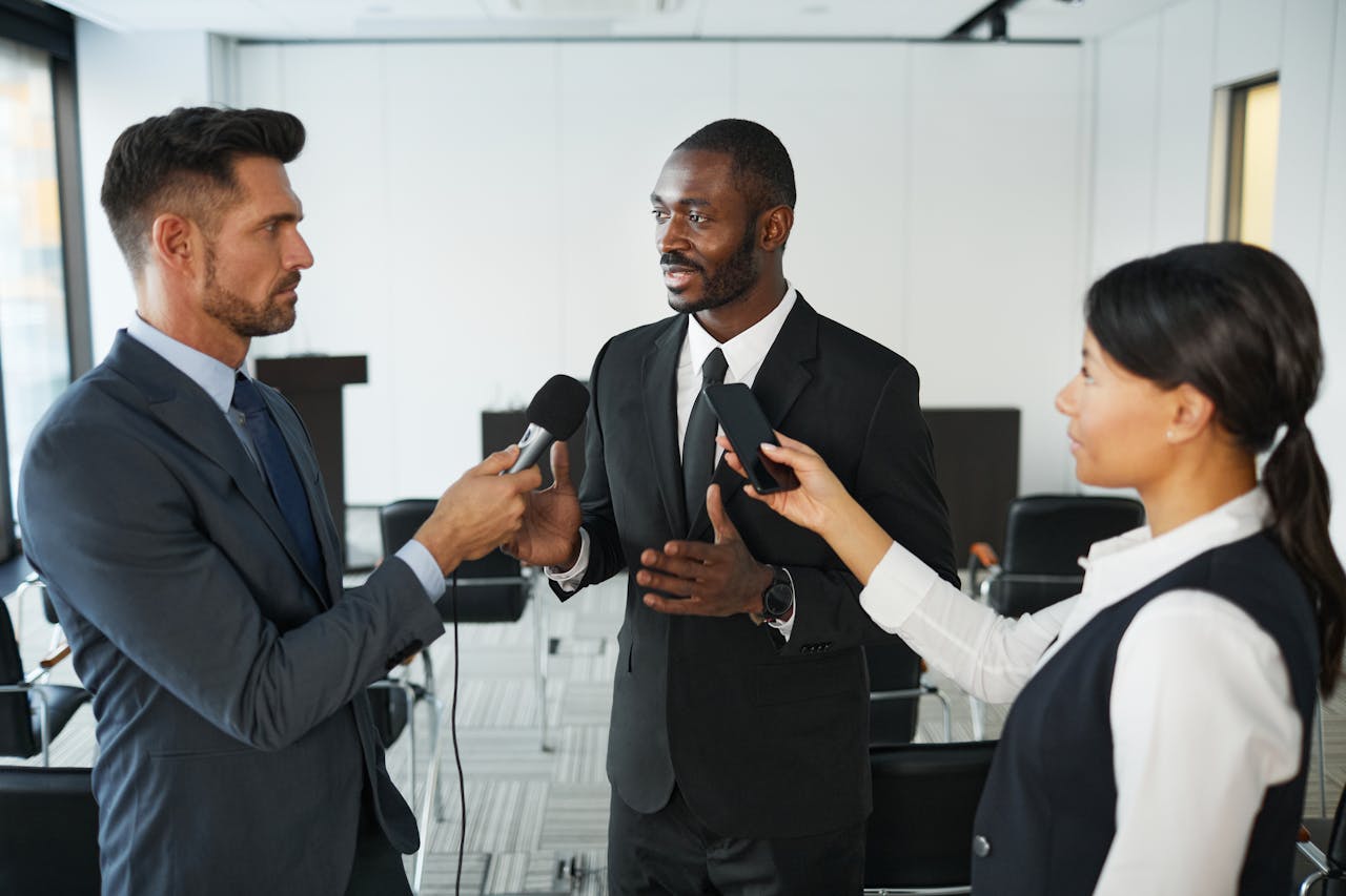 services-03 African American businessman in formal attire being interviewed by journalists with microphones in a conference room.